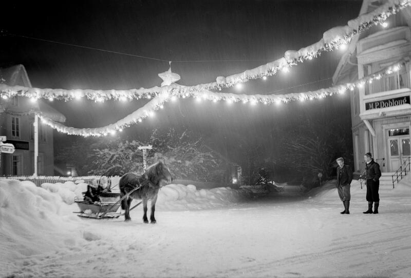 Svart-hvitt bilde av to lyslenker som henger i kryss over snødekket gate, hest med slede til venstre i bildet og to mennesker foran et hus til høyre.