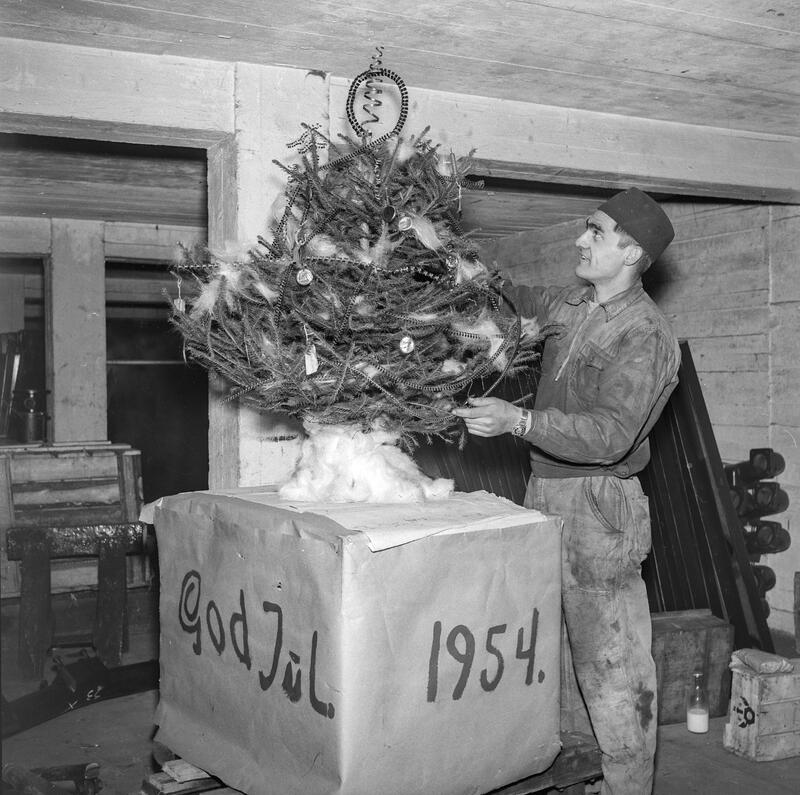 Svart-hvitt bilde av lite juletre som står på en firkantet boks påskrevet "God jul 1954". Til høyre for treet står en mann i kjeledress. Juletreet er pyntet med metallspiraler mm og befinner seg i Hamar jernstøperis lokaler.