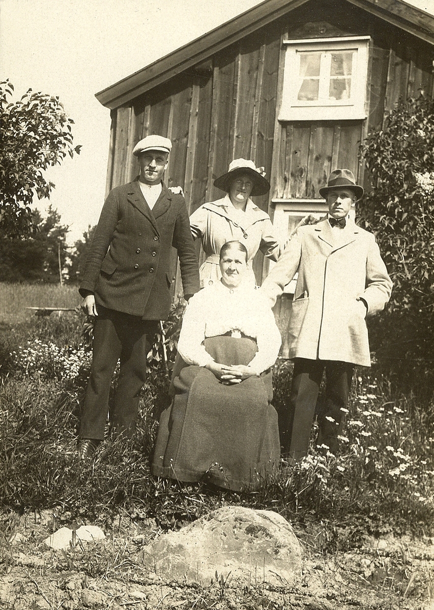 En äldre kvinna sitter framför ett bostadshus, omgiven av sina tre barn.
