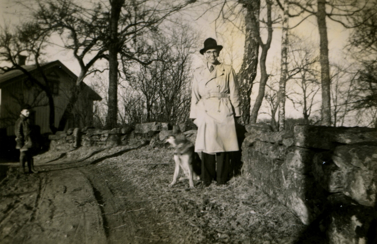En okänd man och en hund står utomhus vid en stenmur i Sporred, okänt årtal. I bakgrunden till vänster står ett barn framför ett hus.
