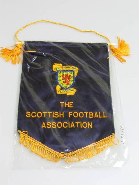 Vimpel från The Scottish Football Association. Vimpeln är i mörkblått siden. På vimpeln finns föreningens logotyp, ett lejon omgiven av tistel i en sköld. Text på vimpeln lyder: "The Scottish Football Association" två gånger. Längst ner hänger en gul fransrad. Vimpeln är monterad på en plaststav som hänger i ett gult snöre som avslutas med tofs på vardera sida. På baksidan står det "Trophy Centre, 28 Kilmarnock RD, Glasgow S41, Tel. 041-6493843". Vimpeln är inplastad.
Vimpeln kommer från Fritidsförvaltningen i Landskrona och lämnades över till museet i samband med Fritidsförvaltningens sammanslagning med Kulturförvaltningen 2005.