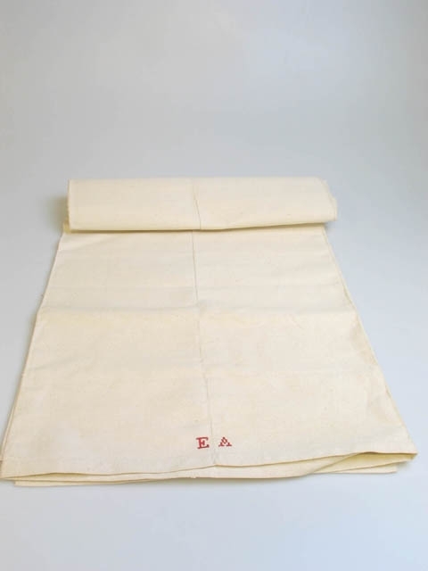 Grovt linnelakan, ihopsytt av två delar. Längst ner monogrammet "EA" broderat i rött med korsstygn. Elin Andersson (1874-1929) var en av systrarna Andersson på Asmundtorp 7 och 9 (Utställningen Systrarna från Asmundtorp). Fyra av de sex systrarna gifte sig inte och deras hemgifter, som bestod av nio kistor med egentillverkade textilier, blev inte använda.