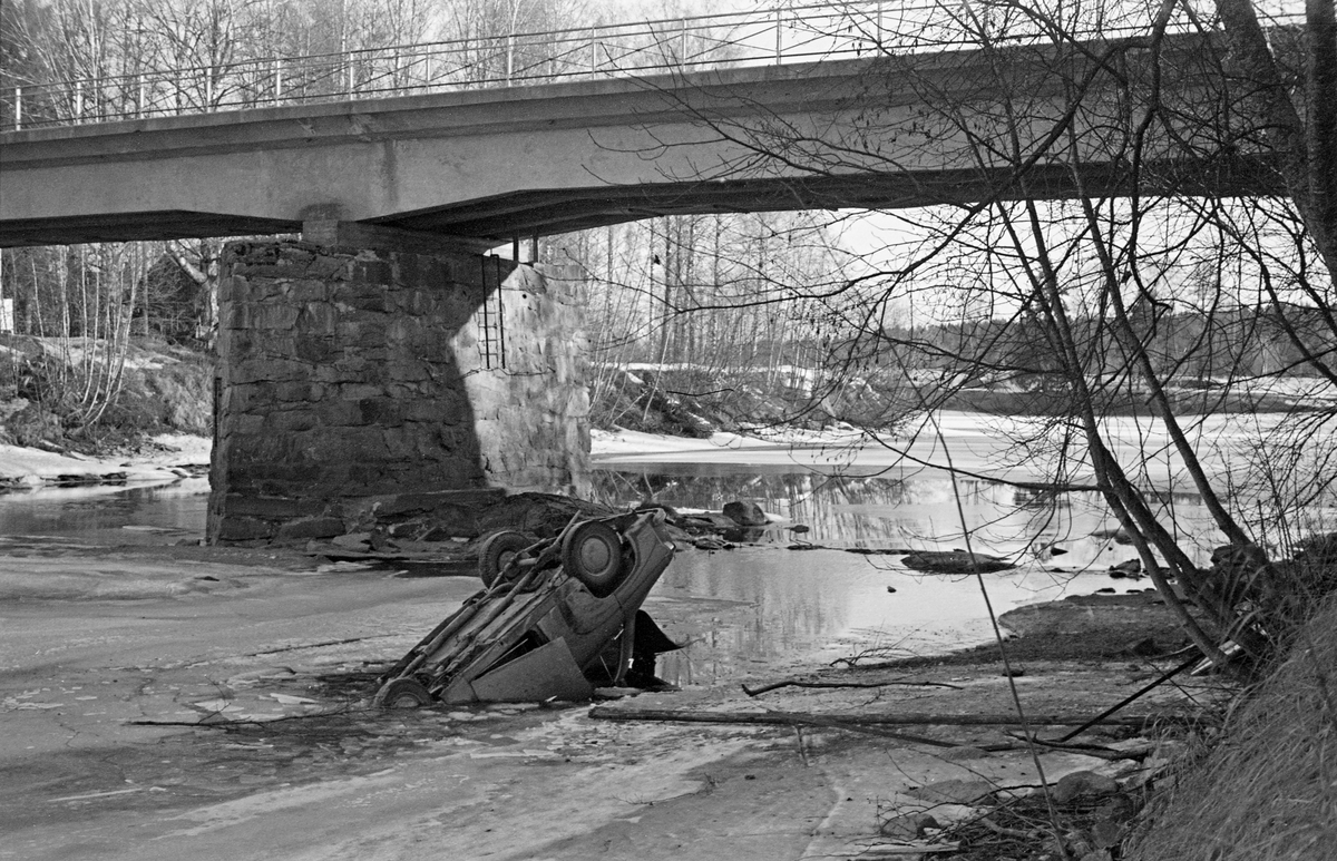 Bil, liggende på taket i den islagte og vintergrunne Oppstadåa, under Osbrua i Sør-Odal i Hedmark etter ei trafikkulykke som skjedde natta mellom 15. og 16. april 1962. Et avisoppslag om ulykka er gjengitt under fanen «Opplysninger».