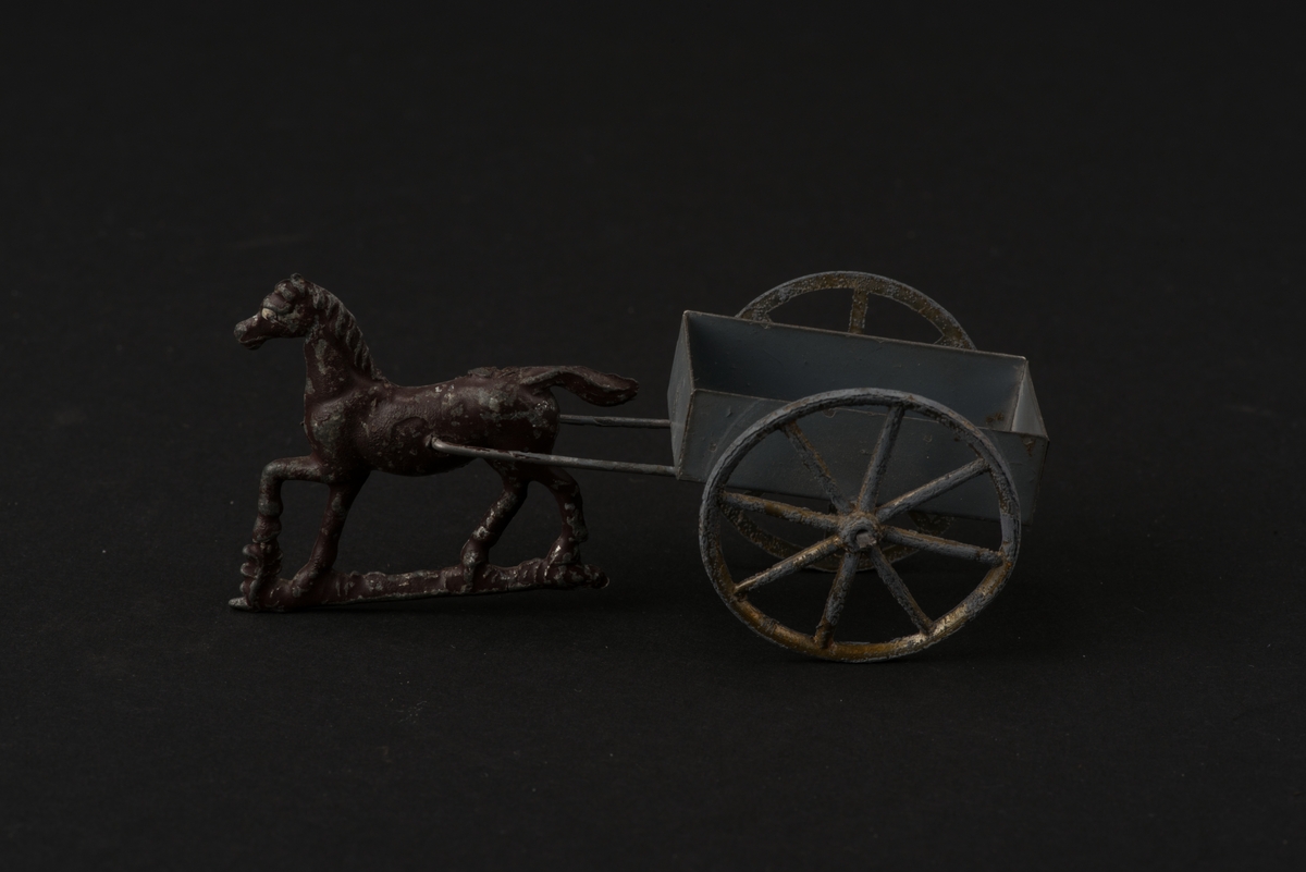 Leksak i form av häst och enaxlad vagn. 
Hästen är gjuten i tenn och brunmålad. Vagnens flak är tillverkat av järnplåt. Flaket och skaklarna är gråmålade. Hjulen var ursprungligen förgyllda.