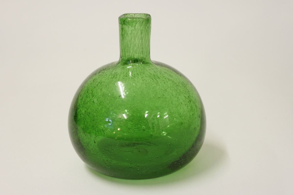 Glasflaska använt i bikupa för socker och vatten. Bullig grön glasflaska med ena sidan slät.