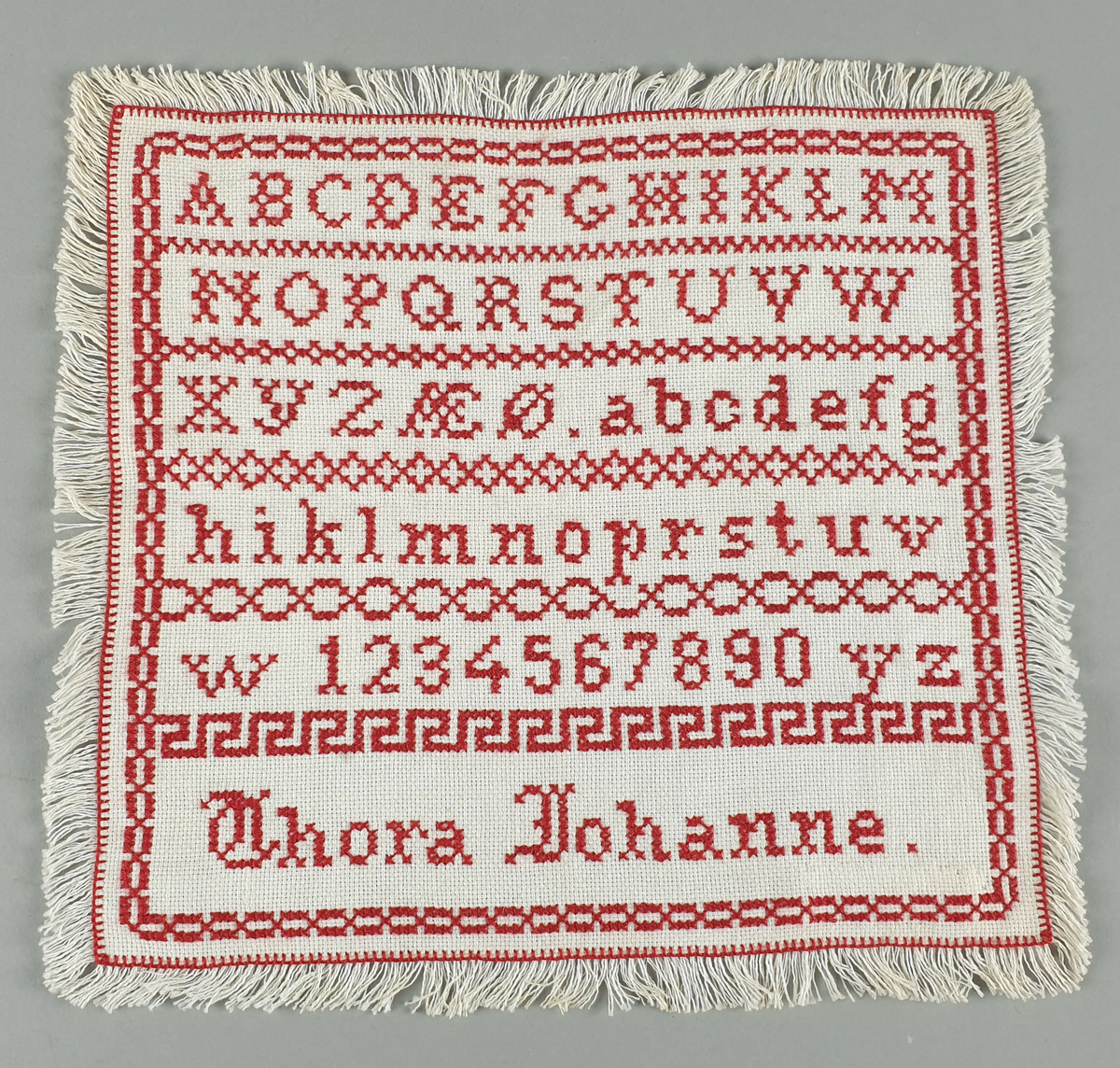 Hvit navneduk i bomull, med rødt broderi av alfabet, tall og egennavn. Border mellom hver linje.