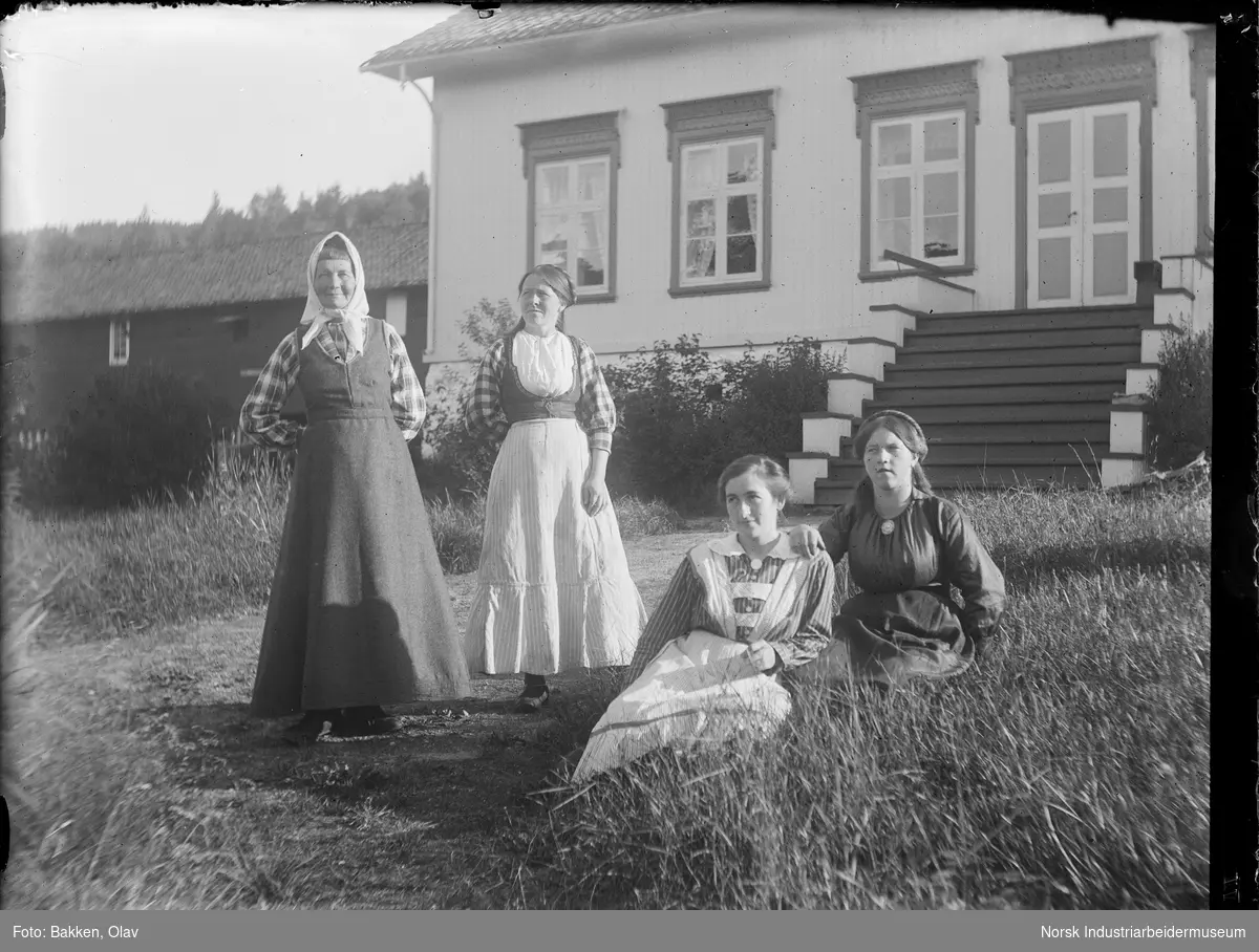 Fire kvinner stående og sittende på gressbakke foran bolighus med trapp. Driftsbygning i bakgrunnen.