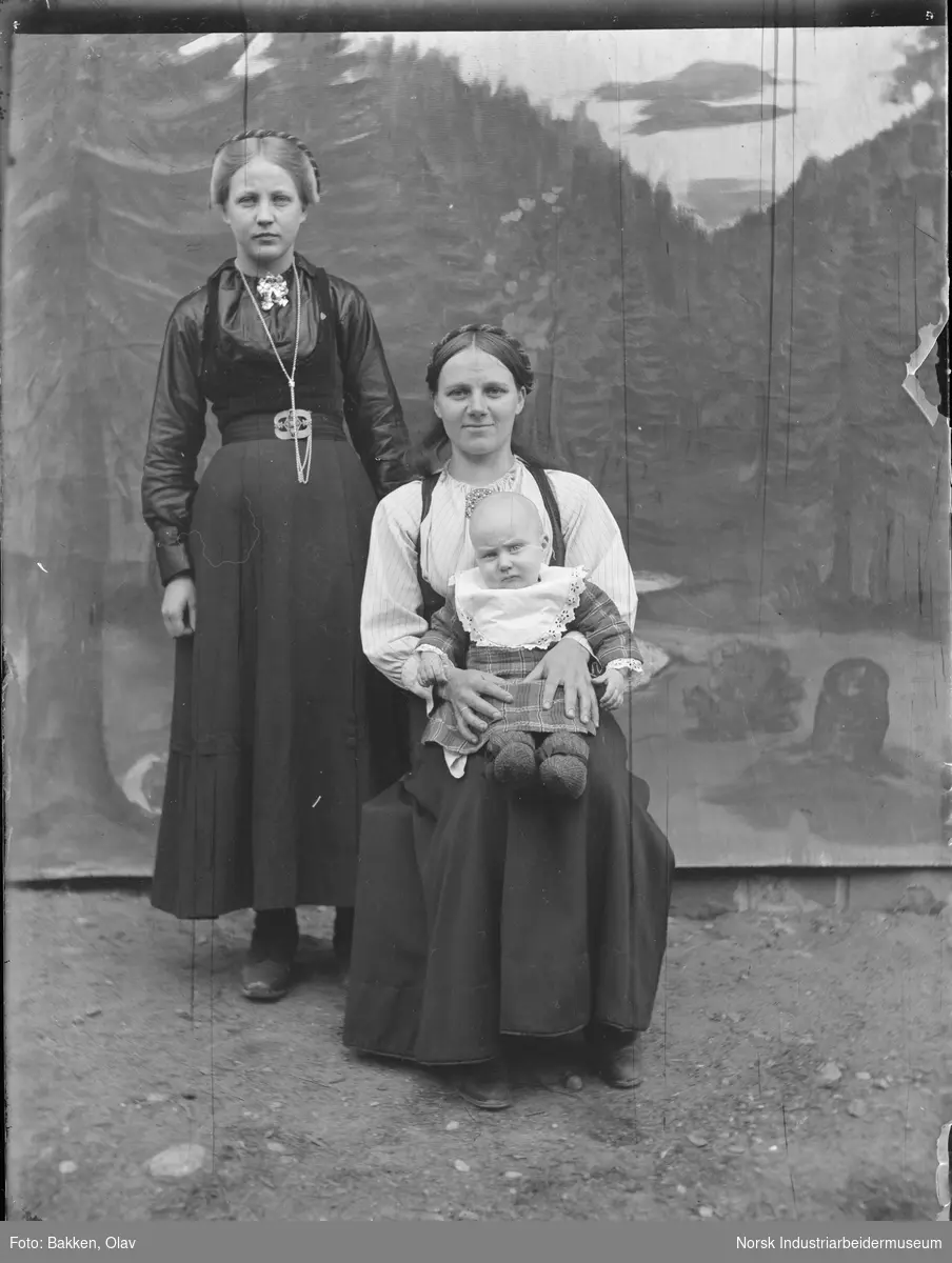 To kvinner og ett spedbarn foran malt fotobakgrunn.