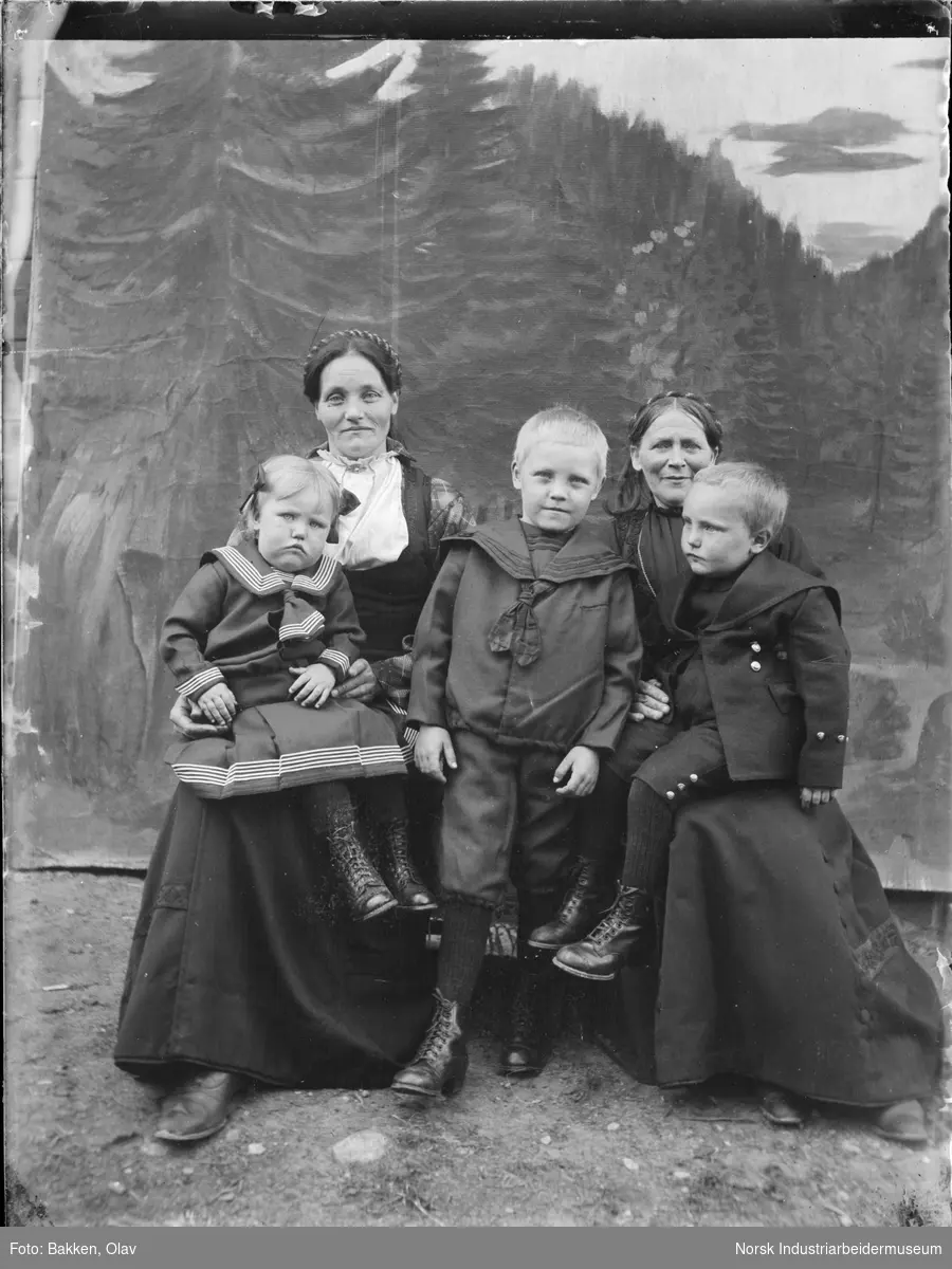 To kvinner og tre barn sittende foran malt bakgrunn. To av barna sitter på kvinnenes fang, et barn står i midten. Alle kledd i pentøy.