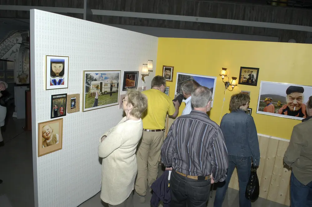 Personer ser på utstillingen. Det henger fotografier og andre bilder på en hvit og en gul vegg.