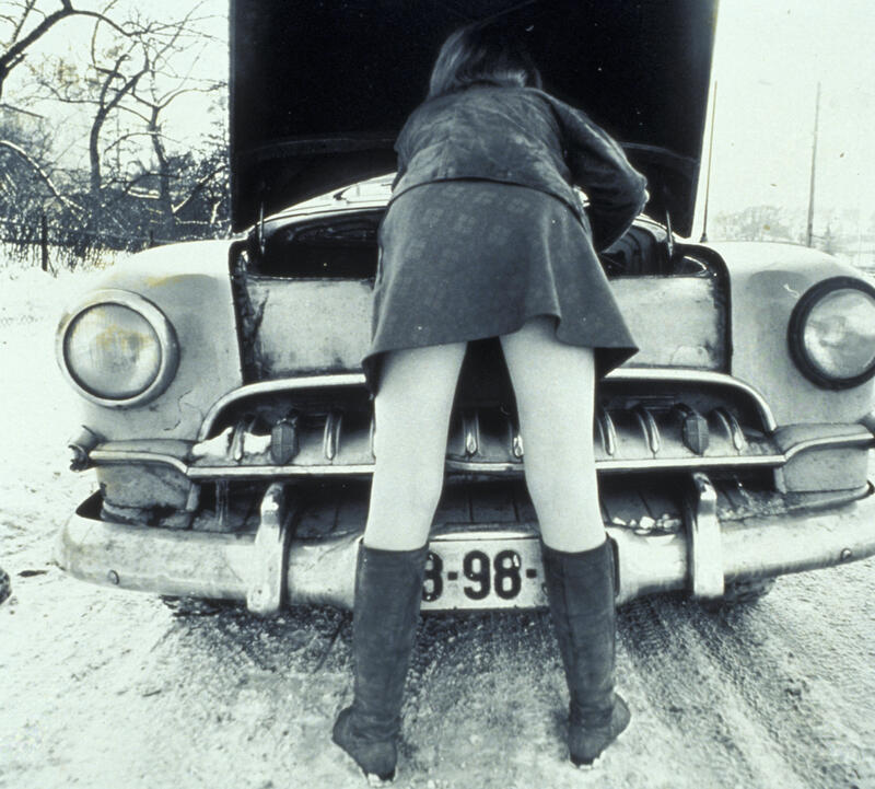 Fotografi av bil med panseret åpent. En kvinne lener seg over og ser ned på motoren.