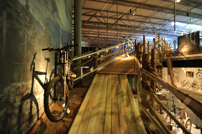 Oversiktbilde av utstillingen Trill rundt. En sykkel står langs en vegg ved starten av en bru.I bakgrunnen ser man flere sykler.