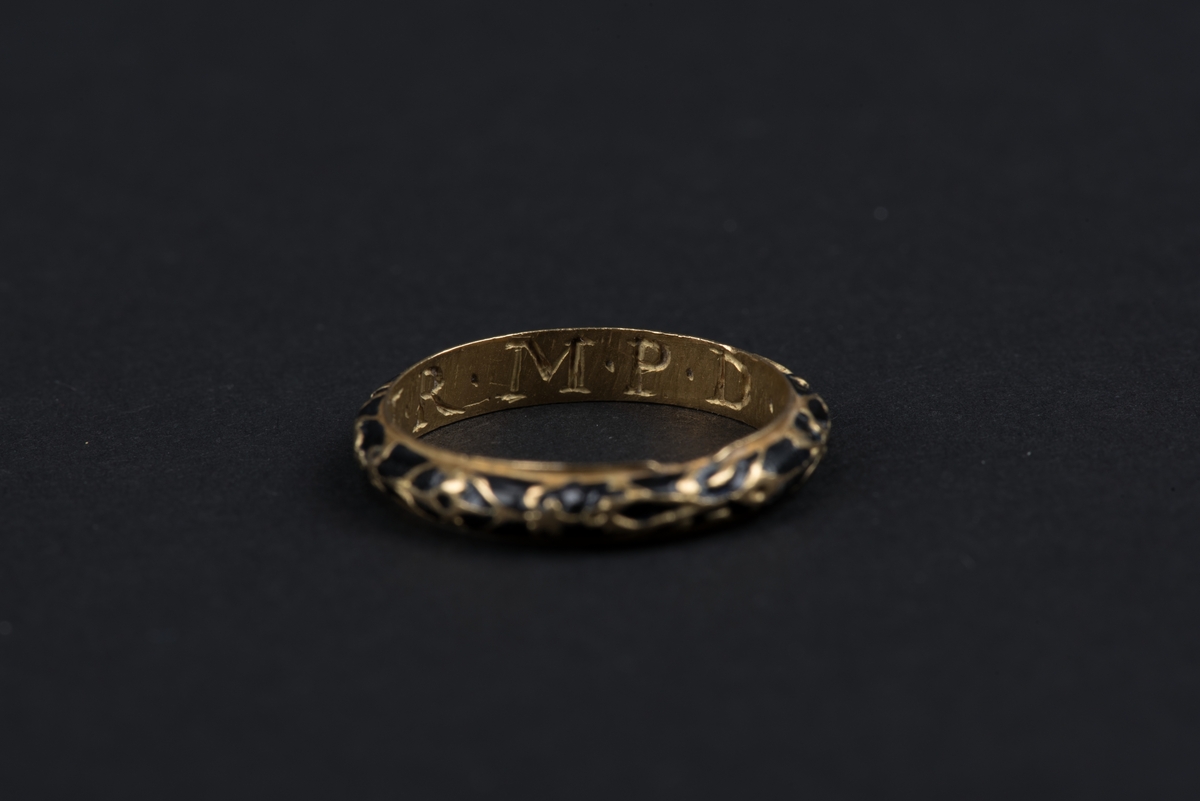 Fingerring av guld med inlagd svart emalj. 
Ringens insida har ingraverade initialer, som indikerar att ringen använts som förlovningsring.