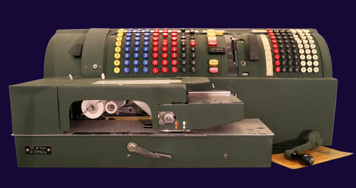 Elektrisk räknemaskin i grön metall. Maskinen står på ett bord med hjul. Siffror och ord på knappar i bakelit i vitt, blått, rött och svart. 
Längst fram på maskinen en tryckdel för att trycka belopp i en bankbok.