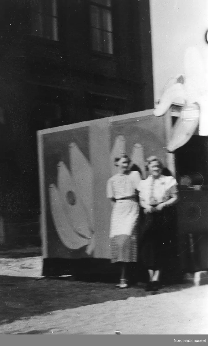 Tordis og Anna Kristine Vatten, datter til Helga og Ole Vatten, foran banankiosken i Bodø. Personene står foran et stort skilt med bilde av bananer. Bygning i bakgrunnen.