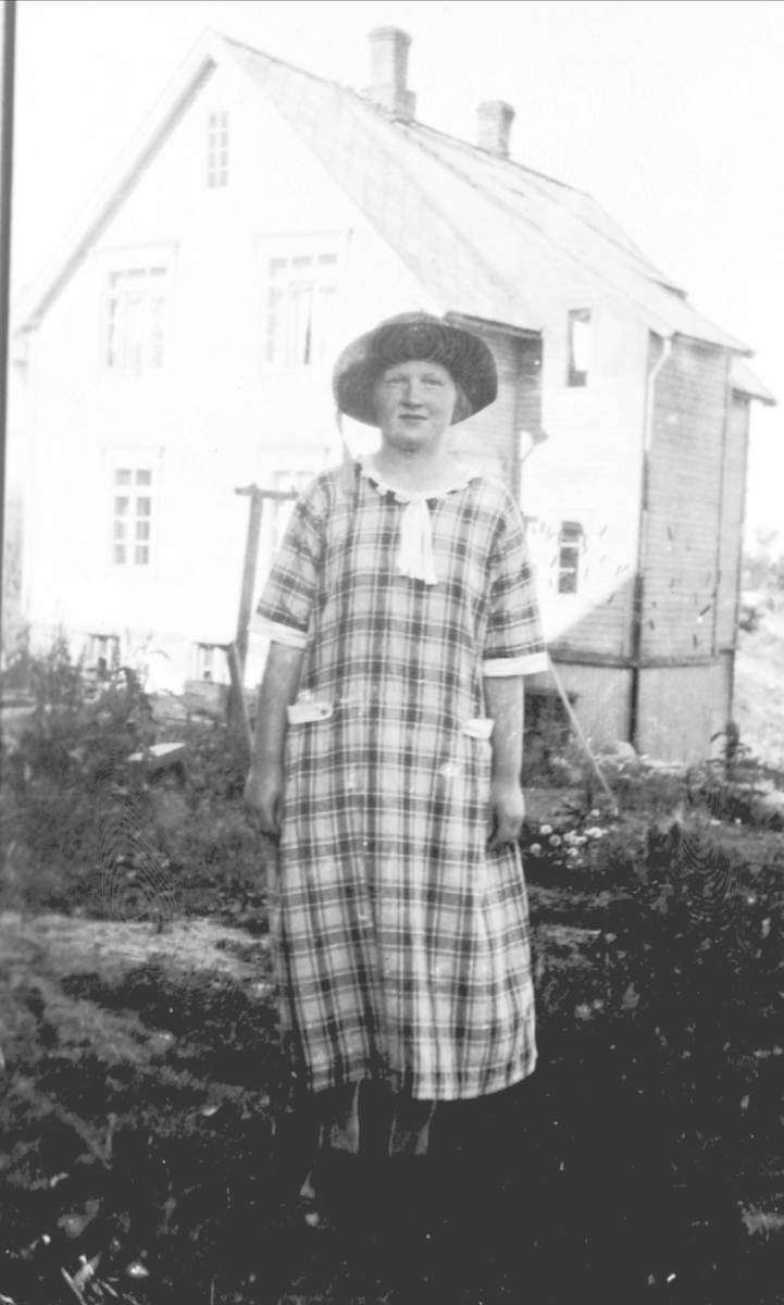 Ung kvinne i sid kjole og hatt, fotgrafert ute foran et hus.