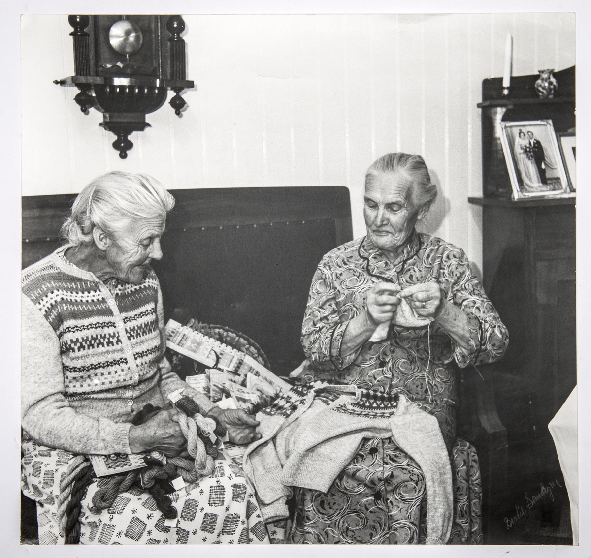 Alma Olsson och Maria Johansson sitter bredvid varandra i en soffa i Marias finrum. Alma har en kofta från Bohus Stickning, Blå Randen - formgivare Anna-Lisa Mannheimer Lunn - och Maria stickar på en kofta till samma företag. Mellan sig har de garner och mönsterdiagram till olika mönster som ingick i Bohus Sticknings kollektioner. Båda stickade åt Bohus Stickning, bilden är tagen inför Marias 80-årsdag och publicerades i Strömstads tidning 16 november 1957.