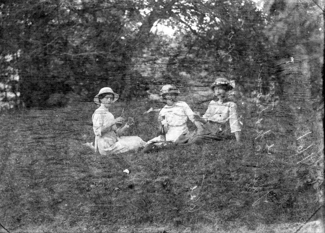 Tre unga kvinnor med hattar sitter på en gräsmatta. De har dricksglas i händer. En mörk flaska står på en bricka. Kvinnan till vänster fyller sitt glas.