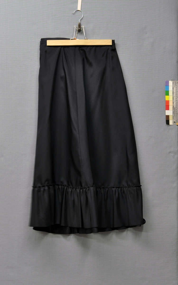 Aftonklänning och skärp i svart sidenorganza med bolero och underkjol.
Klänning (BM63901:1). Avskuren i midjan. Baddräktsliv med påsydd dubbelvikt kant, smockrynkad framtill. Spröt i byst- och sidsömmar. Livet fodrat med svart viskostaft. Rundskuren solfjädersplisserad kjol, blixtlås i vänster sida. Skärp med tygklätt spänne.
Jacka (BM63901:2). Bolerojacka. Framsidan med rundskuren kant och 2 bystinsnitt. Baksidan helskuren med 2 midjeinsnitt. Ståkrage bak, skuren i ett med framstycket. Holkärm. Rullfållad med vändsydda sömmar.
Underkjol (BM63901:3) av 2 våder svart viskostaft med rynkad volang nedtill. 25 mm bred linning, knäppt med 1 knapp. 1 midjeinsnitt i var sida. Storlek 42.