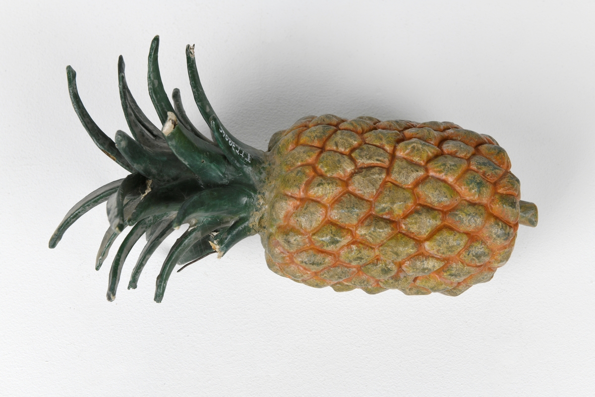 En modell av en ananas støpt av gips med ståltråd i. Ananasen er malt i gulbrunt og oransje. Bladene på toppen er laget av papir og ståltråd (pappmasjé) som er malt grønn. Modellen er vokset (har et lag voks utenpå malingen).