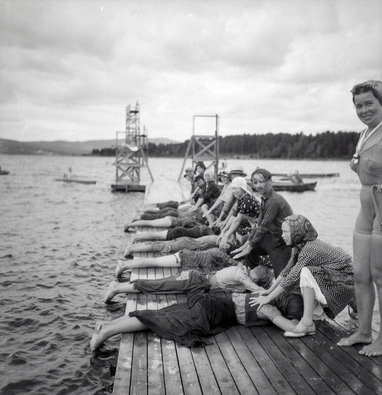 Barn ligger påkledd på magen på en brygge mens de har hver sin medhjelper til å trene på svømmeteknikk.