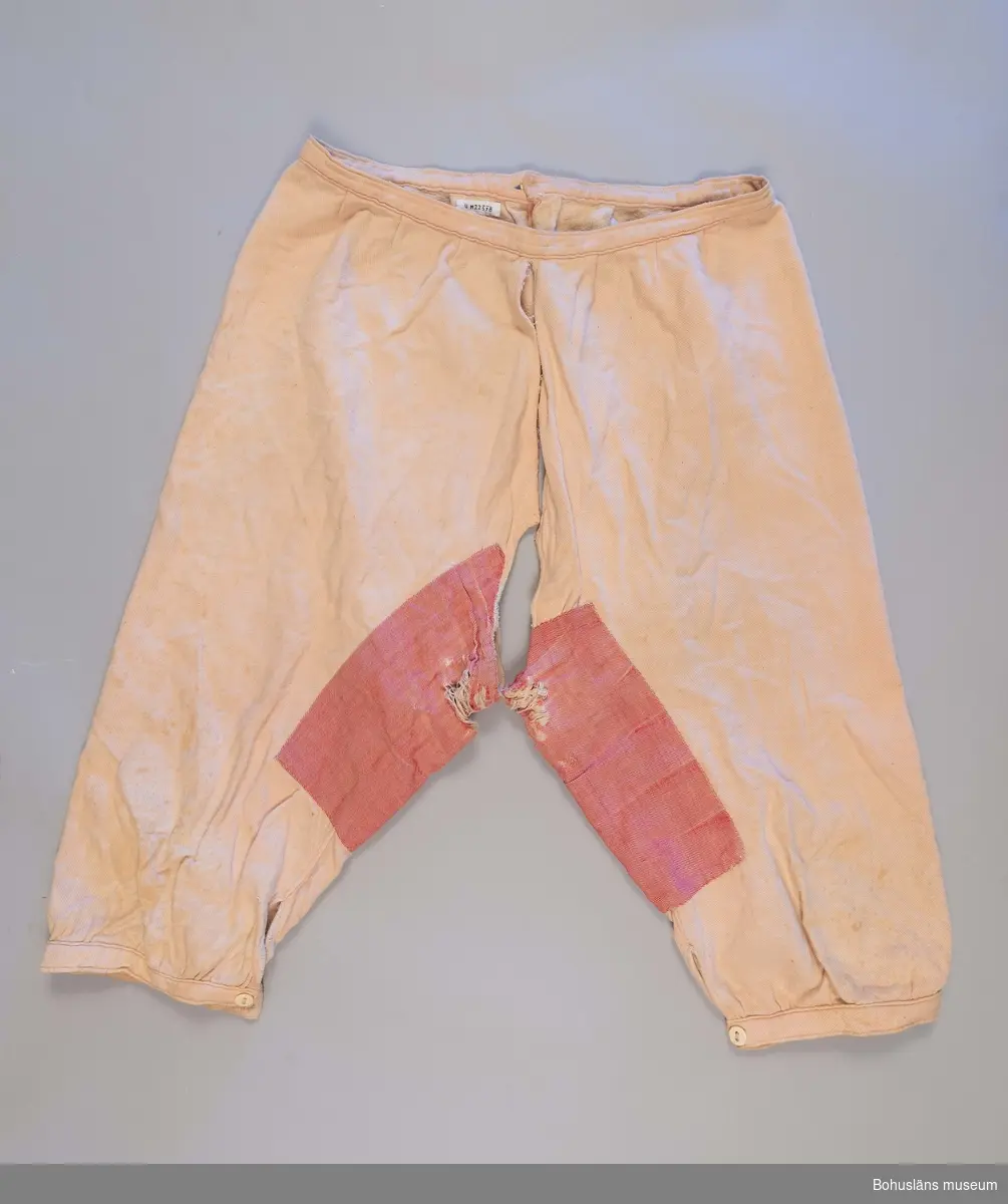 Underbyxor från omkring  1920-1930-talet med sprund i grenen samt knäppning med benknappar vid knäna och linningen. 
Lagade med rött tyg på insidan av byxbenen.