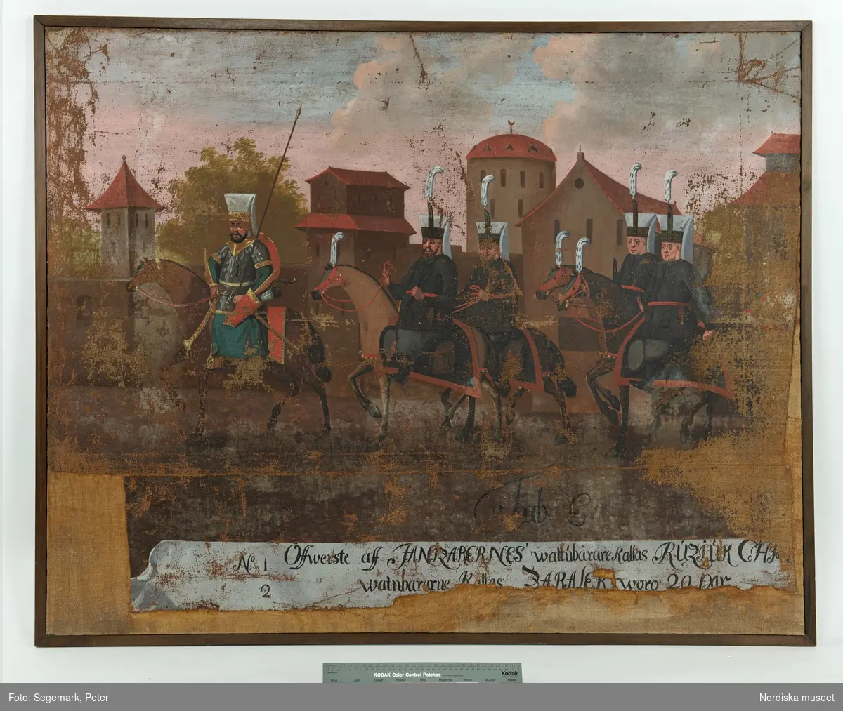 Målning, tavla, Tabula E, processionssvit, en av totalt 20 målningar föreställande en procession genom Konstantinopel (nuvarande Istanbul) med den turkiske sultanen Mehmet IV och hans hov på väg till den årliga höstjakten den 24 september 1657.

Stadsmotiv med byggnader i bakgrunden, procession från höger till vänster i bild. Längst fram en ryttare med hög huvudbonad, pilkoger vid midjan hållande en lång käpp vertikalt. Efter honom fyraridande män med blå rockar och grå byxor. Mannen längst fram markerad med siffran "1", en av de efterföljande männen med siffran "2". Under processionen texten "Tab.E." Längst ned textremsa med texten: "No  1 Öffwerste aff ZANIZARERNES wattubärare Kallas KUZJUK CHJ---
No  2  Watnbärarne Kallas ZAKALER woro 20 par".

Anm.
Skador längs kortsidor och nedre långsida, se konserveringsprotokoll.
/Maria Maxén 2014-08-15