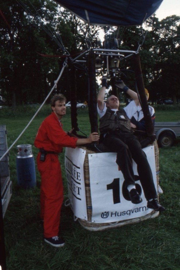 Två personer står och håller i korgen till en luftballong. En person sitter på kanten till ballongkorgen och ordnar med dess brännare. På korgen står det "Andréemuseet" och "16".