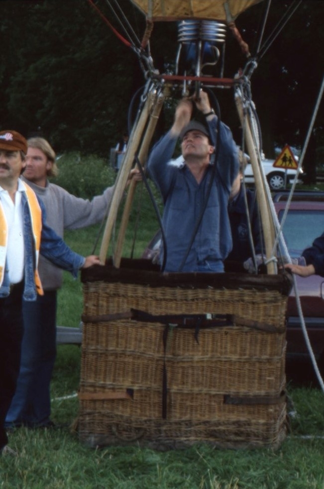En man står i en ballongkorg och pysslar med dess brännare. Utanför till vänster står två personer och håller i ballongkorgen.