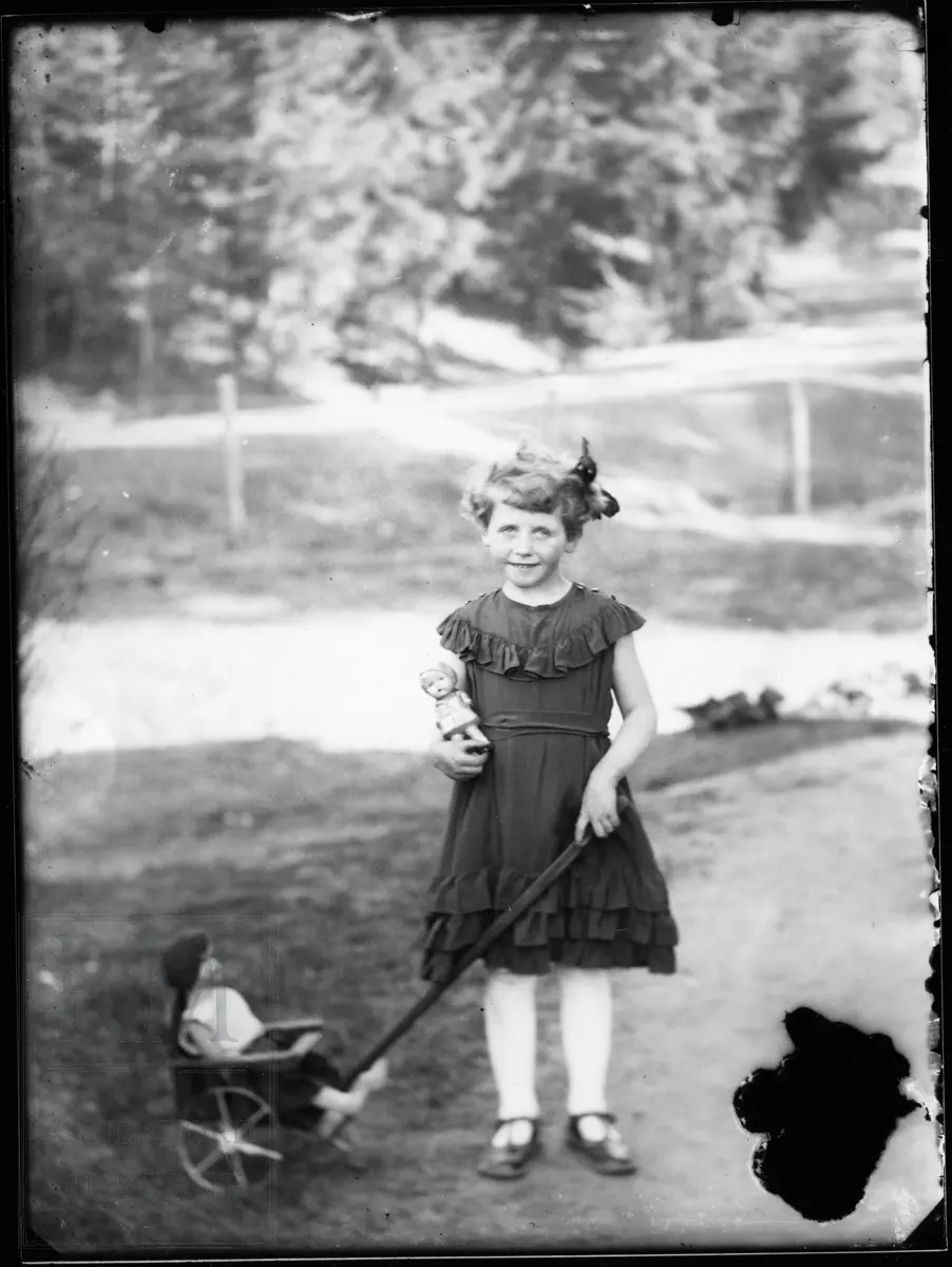 En lita jente med kjole og sløyfe i håret, står og holder ei lita lekevogn med ei dukke i.