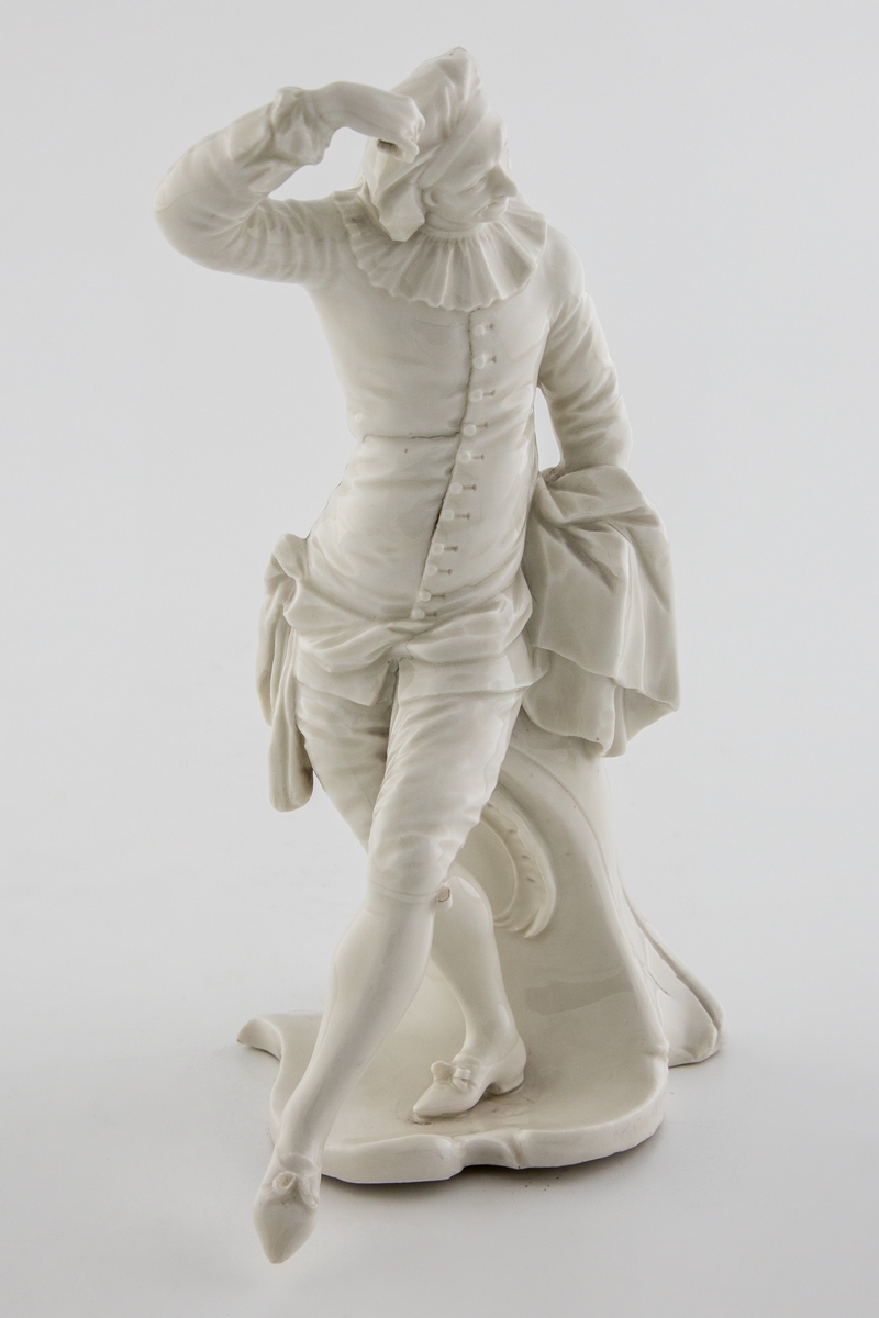 Hvitglasert, umalt porselensfigur av en dansende mann i 1700-talls klær som støttes av et postament med rokokko-trekk. Med høyre hånd tar han på sin lue og med venstre hånd holder han kappen åpen. Figuren beskrives både som harlekin, og identifiseres som scaramuzzio (også: scaramuccia, scaramouche eller scaramuz), en karakter fra commedia dell´arte som oftest blir fremstilt i svart spansk drakt.