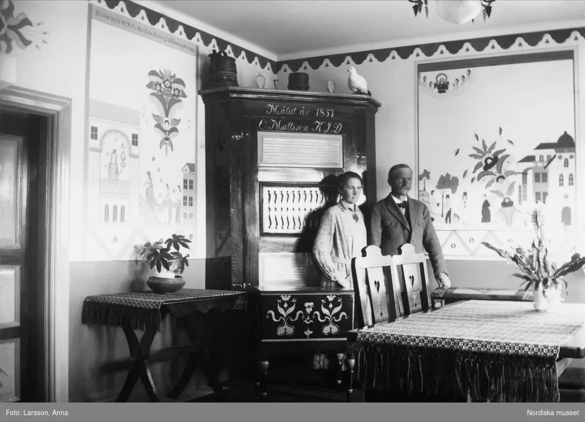 Paret Almgren i sitt hem, stående framför väggskåp med kurbitsmålningar på väggarna