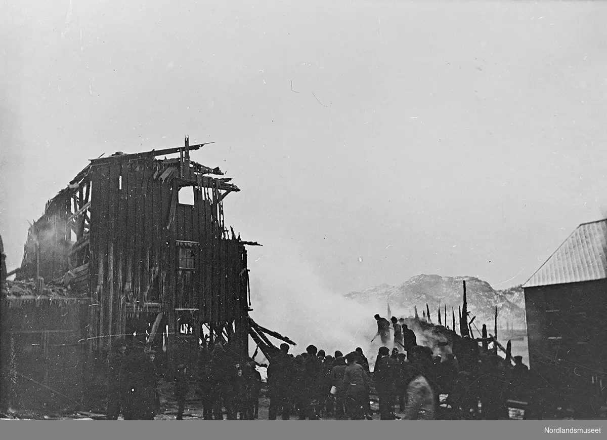 Postkort. Bodø, kullageret brenner. Motivet viser brannherjet hus. Mange mennesker er samlet nede på kaia.