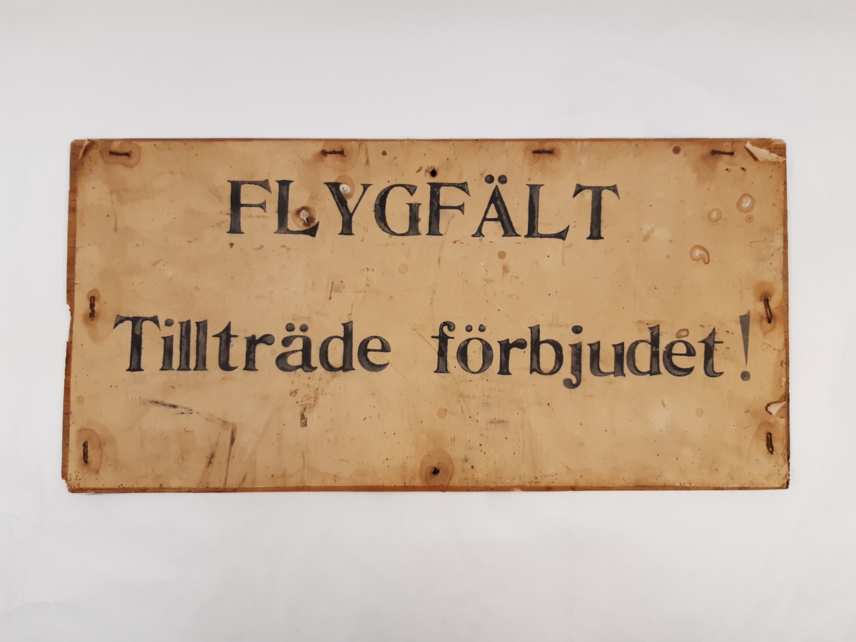 Förbudsskylt med texten "FLYGFÄLT", "Tillträde förbjudet". Skylten består av en spånskiva med på fronten häftat papper.