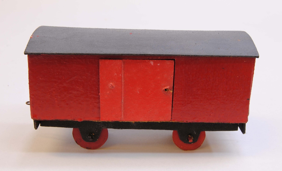 Röd godsvagn av papp.
Delarna är limmade eller sammanfogade med rött lack. Hjulen är gjorda av papp och hjulaxlarna av fyrkantiga träpinnar, troligtvis tändstickor. Vagnskorgen är målad röd med svart underrede och röda hjul. På ena långsidan finns en dörr. Taket är lätt välvt och mörkgrått.
Under vagnen är datumet "28/12 1918" handskrivet. På kortsidorna av vagnen finns böjd ståltråd, en kork och en ögla för att koppla samman vagnen med andra vagnar eller lok.