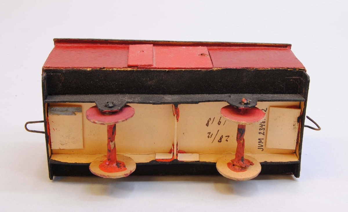 Röd godsvagn av papp.
Delarna är limmade eller sammanfogade med rött lack. Hjulen är gjorda av papp och hjulaxlarna av fyrkantiga träpinnar, troligtvis tändstickor. Vagnskorgen är målad röd med svart underrede och röda hjul. På ena långsidan finns en dörr. Taket är lätt välvt och mörkgrått.
Under vagnen är datumet "28/12 1918" handskrivet. På kortsidorna av vagnen finns böjd ståltråd, en kork och en ögla för att koppla samman vagnen med andra vagnar eller lok.