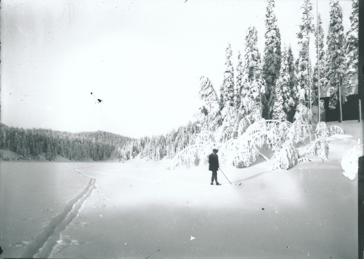 Mann på ski på innsjø. Skispor er synlig i snøen. Til høyre er en knaus med skog og hytte.