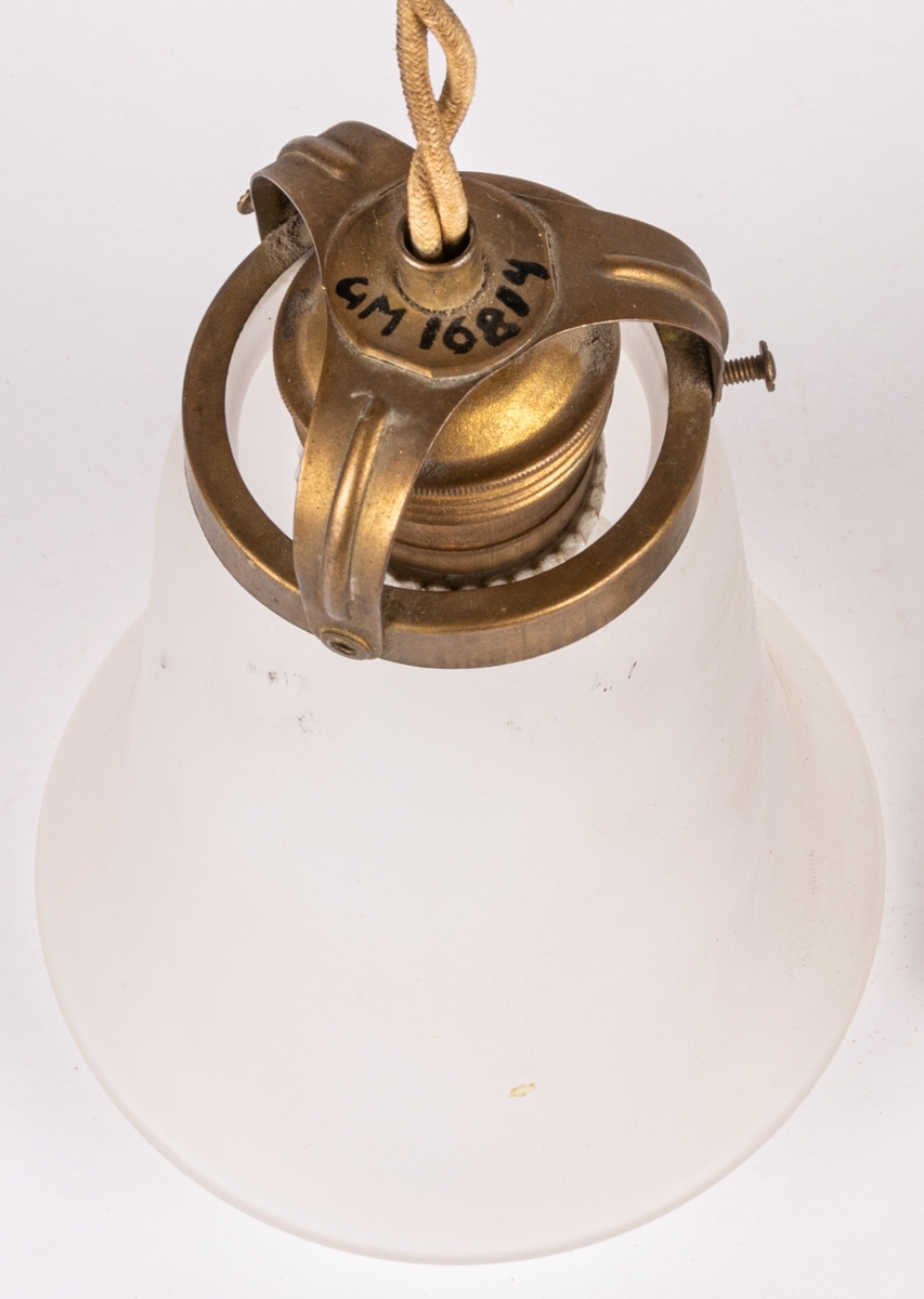 Taklampa, glas och mässing. Slät klockformad kupa av matt glas med invikt kant. Kupan sitter fast i en mässingsbehållare som hänger i en snodd från en rund takskiva i mässing.