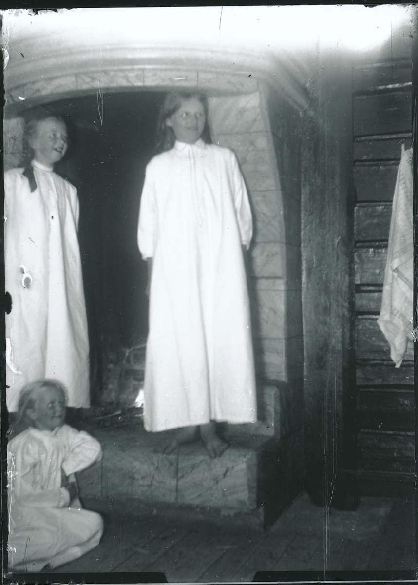 Tre jenter i lange nattkjoler som står eller sitter foran peis i hytteinteriør.