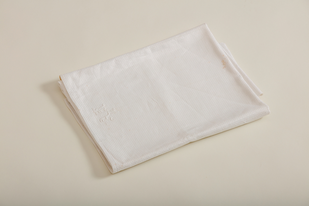 Håndkle av hvit lin med mønstereffekt i vevningen. Maskinsydd fold i hver ende. Hvitt håndbrodert monogram i plattsøm. Påsydd hank av bomullsbånd.
