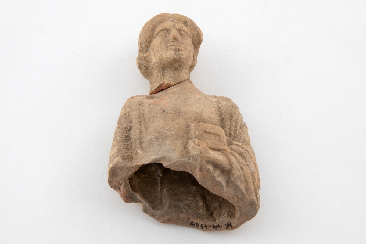 Fragment av figurin i terrakotta, øvre halvdel av kroppen er bevart. Fremstiller en kvinne iført drakt med oppsatt hår. Hun holder en eske i venstre arm. Figurinen har brudd ved halsen, og er senere limt. Lyst, rødlig gods, med grålige partier.
