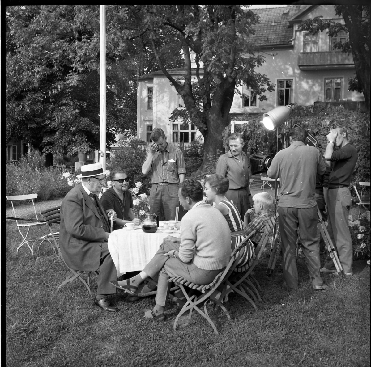 En man, en kvinna och två pojkar sitter vid ett trädgårdsbord i en trädgård. Framför dem sitter två herrar. Ståendes runt bordet befinner sig fyra personer. En filmar de sittandes.