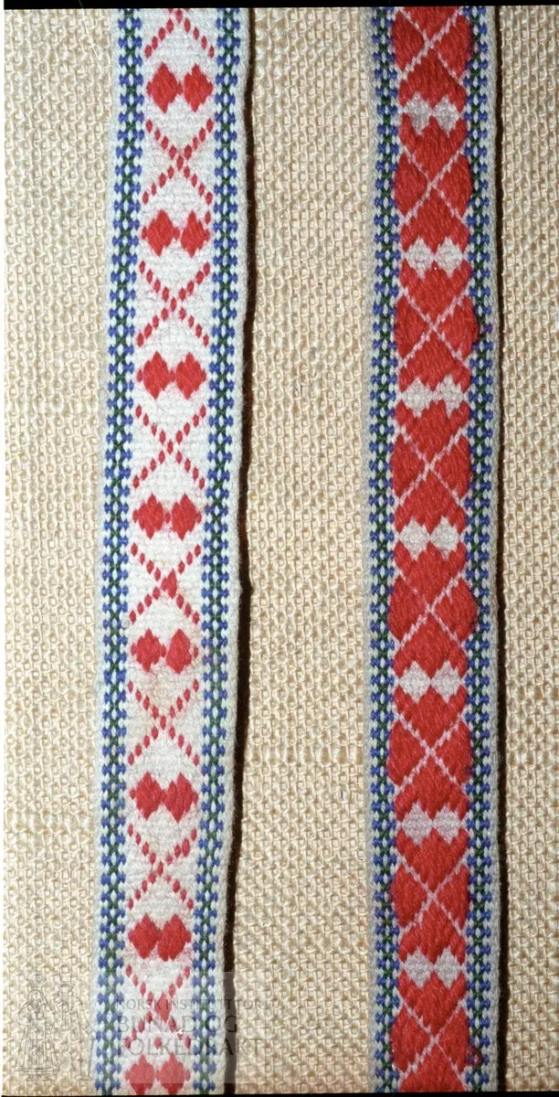Grindvovne band med plukka mønster. Kvitt bomullsgarn, mønster i ullgarn, raudt, blått og grønt. Bredde:  3 cm, lengde:  85 cm.