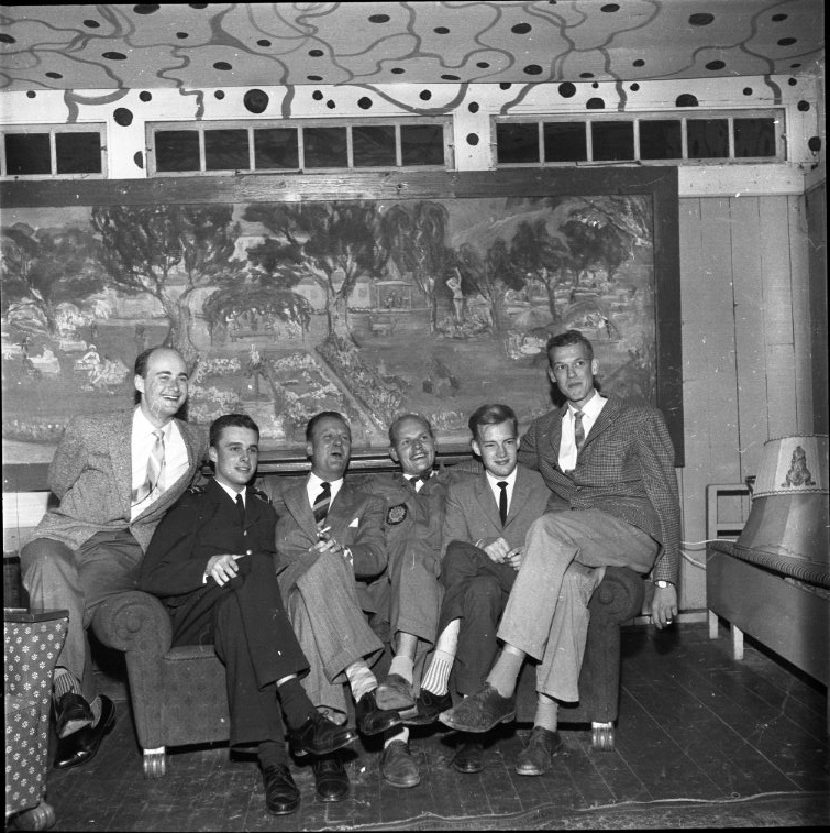 Sex herrar sitter i en soffa. På väggen bakom dem hänger Karin Stacklebergs målning "Ribbagårdens trädgård", daterad 1935.