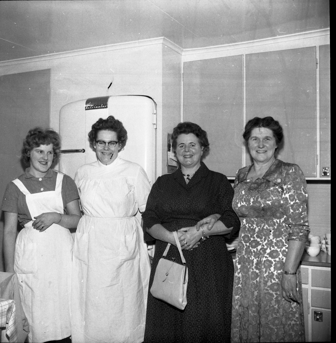 Fyra damer står uppradade i ett kök. Bakom sig har de en frys av märket Kelvinator. De har kortklippta, lockiga frisyrer. En kvinna bär ett vitt förkläde och en annan en vit särk.  De andra två är klädda i mönstrade klänningar.