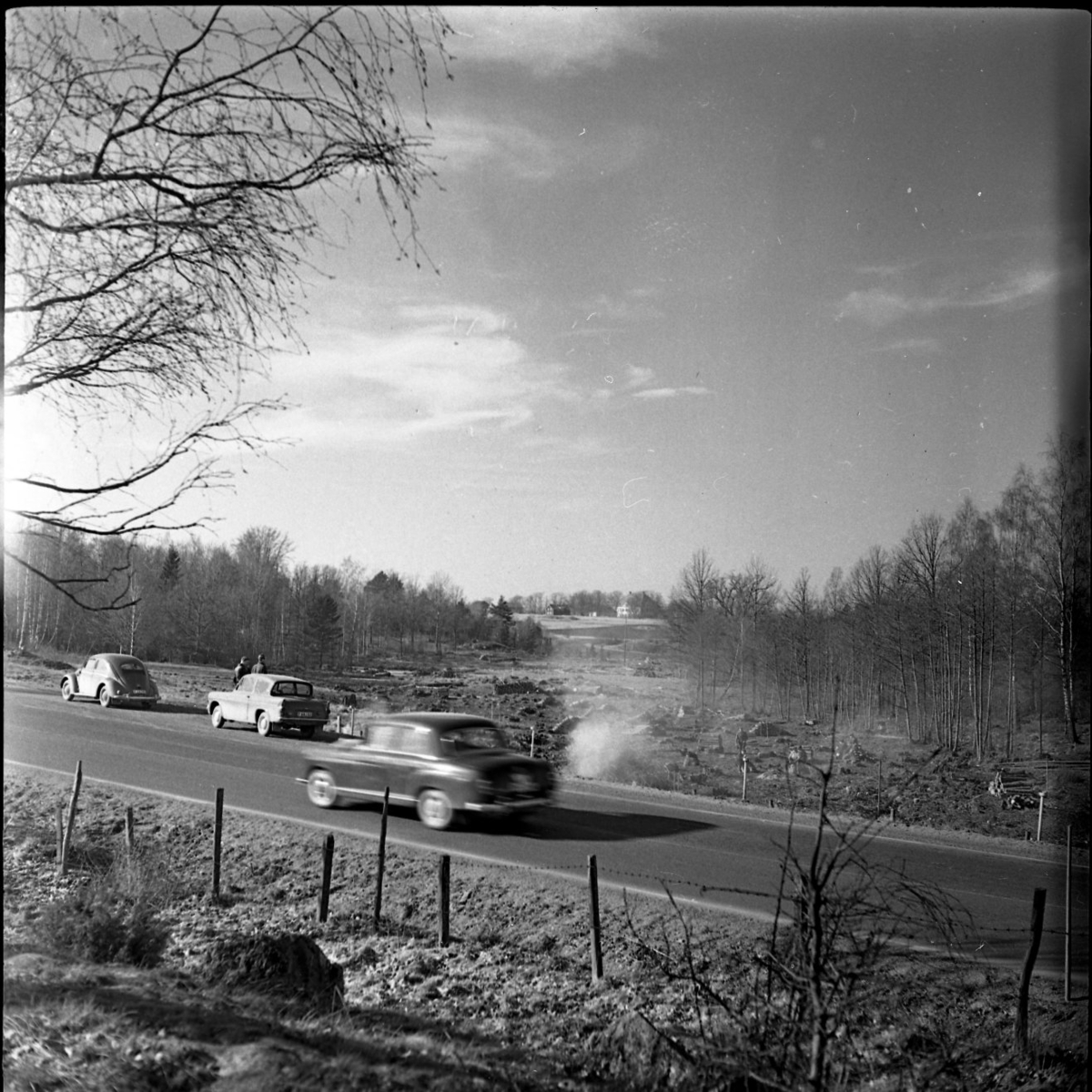 Vy mot rikshuvudväg 1. En bil fotograferas i farten. Bortanför vägen står arbetare och avverkar träd inför motorvägsbygge. I bakgrunden syns Västanå slott med flera byggnader.