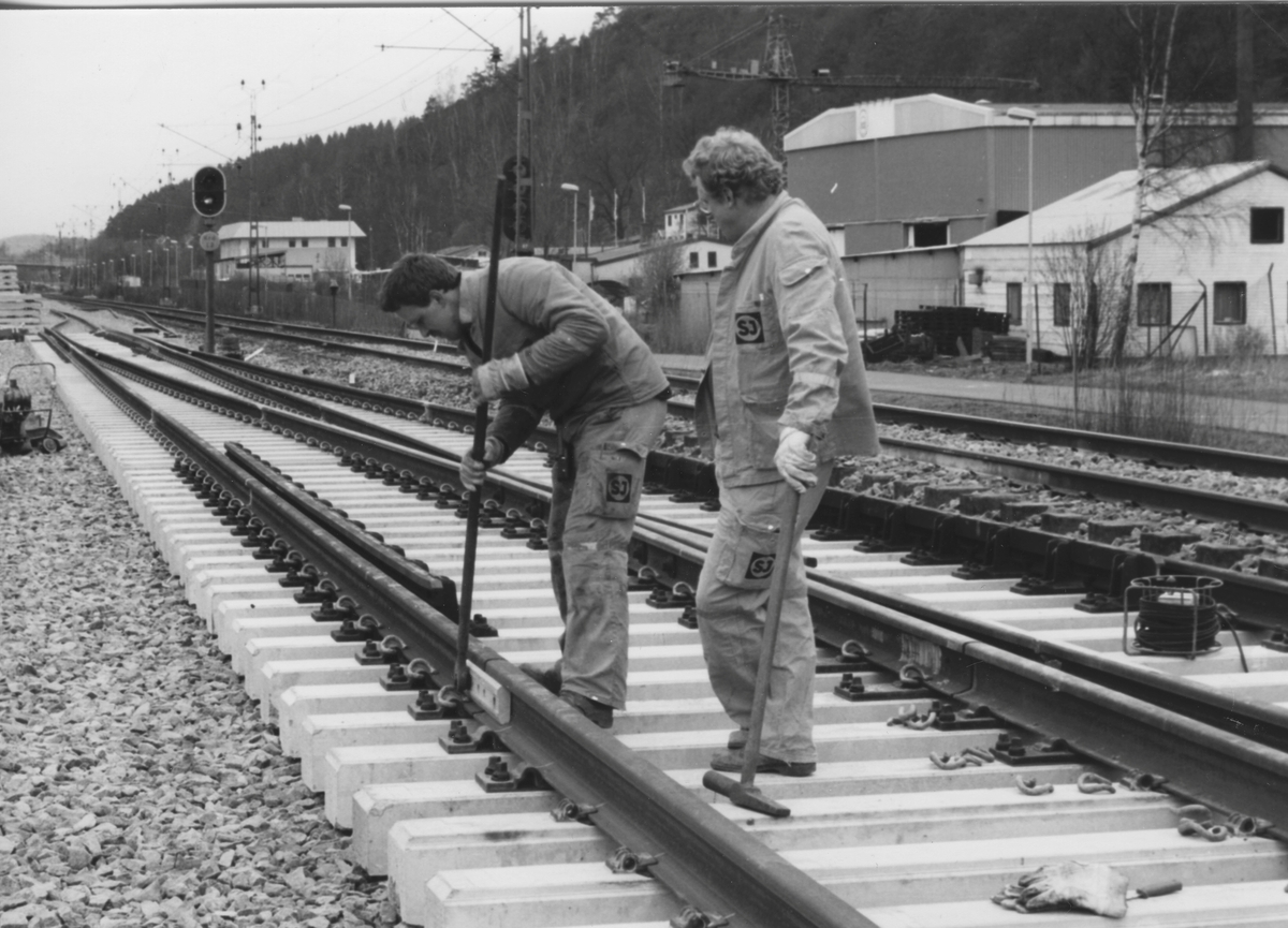 Utbyggnad för pendeltåget 11 april 1990. Två män arbetar med järnvägsspåren vid Kållereds norra infart. Till höger ses fastigheter utefter Gamla Riksvägen.