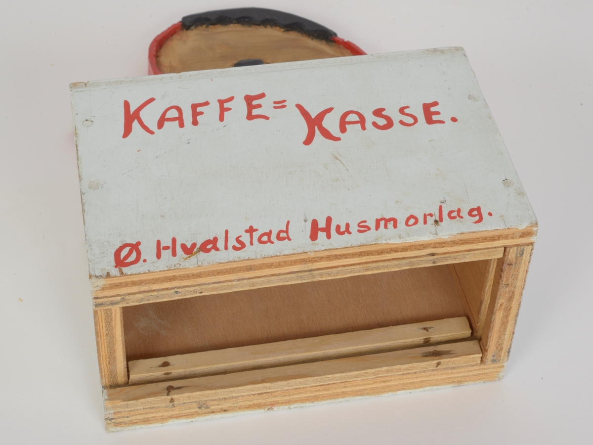 Sparebøsse/kaffekasse i bemalt tre. En hvit rektangulær boks med  påmalt rød skrift; KAFFE=KASSE. Ø.Hvalstad Husmorlag. På oversiden av boksen er det et lite hull med plass til å legge i penger. En utskåret rød kaffekanne over dette.