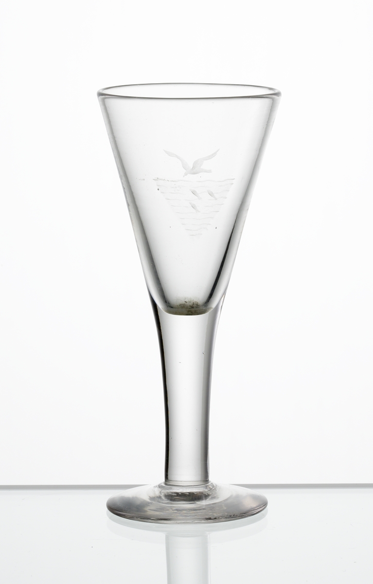 Design: Edward Hald.
Brännvinsglas. Konisk kupa med graverat motiv i form av en mås som flyger över vatten med tre fiskar i.
