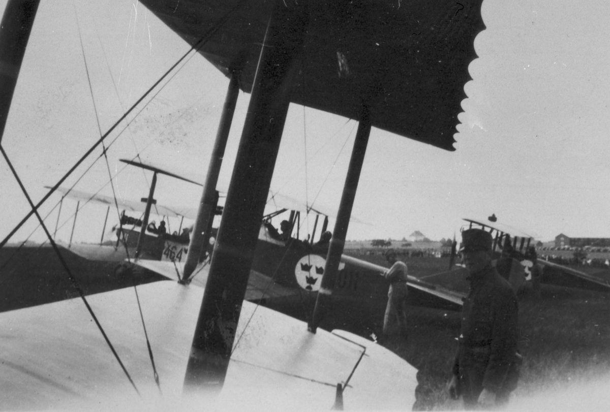 Fyra militära flygplan Albatros B. II står startklara på ett flygfält. Flygförare på plats i flygplanen och andra militärer bredvid. Bild tagen från ett av flygplanen med vinge i förgrunden. Två flygplan märkt nummer 464 samt 011.  Cirka 1926.